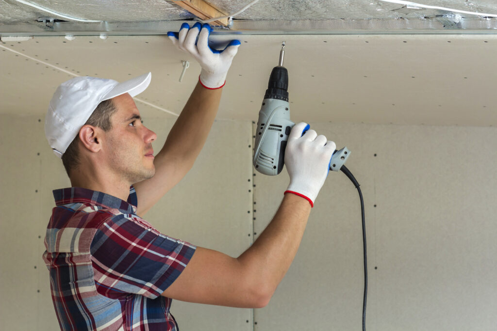 Young man in usual clothing and work gloves fixing drywall suspended ceiling